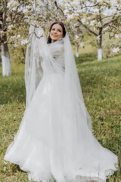 Çok güzel bir gelinin portresi, uzun kıvırcık saçlı, beyaz bir elbise ve çiçek açan bir bahar bahçesinde uzun bir peçe takan. Naarechena ellerini çimenlerin yeşil arka planında tutuyor.