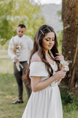 Bir gelin ve damat arkalarında bir ağaçla bir tarlada duruyorlar. Gelin elinde bir çiçek tutuyor.