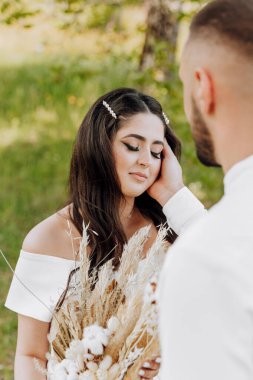 Gelin ve damat bir tarlada duruyorlar, gelin bir buket kuru ot tutuyor. Adam gelinin koluna dokunuyor ve kadın da ona bakıyor. Sahne romantik ve samimi.