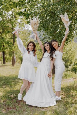 Üç kadın tarlada fotoğraf çektirmek için poz veriyor, biri beyaz elbiseli.