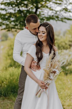 Bir gelin ve damat tarlada sarılıyor. Gelin elinde bir buket çiçek tutuyor.