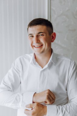 Beyaz gömlekli genç bir adam gülümsüyor ve bir resim için poz veriyor. Beyaz bir gömlek giyiyor ve elleri cebinde.