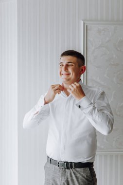 Beyaz gömlekli bir adam giyiniyor ve gülümsüyor. Siyah kuşak ve gri pantolon giyiyor.