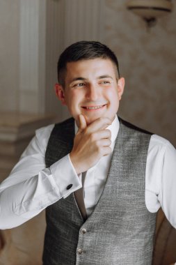 Takım elbiseli ve yelekli bir adam gülümsüyor ve kameraya bakıyor. Kravat ve yelek giyiyor ve gülümsemesi mutlu ya da kendinden emin olduğunu gösteriyor. Profesyonellik ve resmiyet kavramı