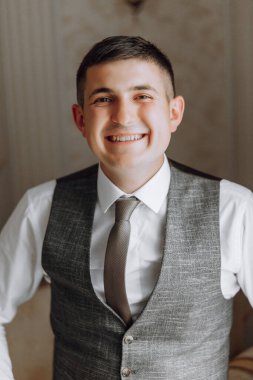 Gri yelek ve kravat takan bir adam gülümsüyor. Yeleğin altına beyaz bir gömlek giyiyor.