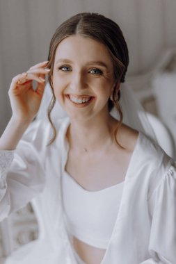 Bir kadın gülümsüyor ve beyaz bir bornoz giyiyor. Bir sandalyede oturuyor ve saçı topuz gibi.