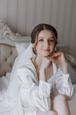 Beyaz elbiseli bir kadın elleri dizlerinin üzerinde bir yatakta oturuyor. Yüzünde bir gülümseme var ve peçe takıyor.