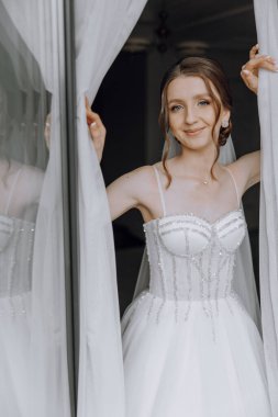 Beyaz elbiseli bir kadın pencerenin önünde duruyor, gülümsüyor. Elbise pullar ve incilerle süslenir, ve kadının saçı topuz şeklinde şekillenir. Zarafet ve mutluluk kavramı