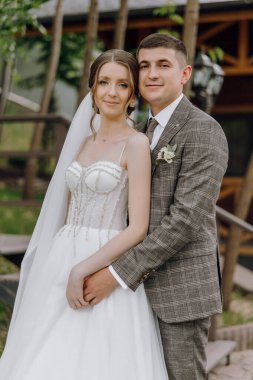 Bir gelin ve damat ahşap bir merdivenin önünde fotoğraf için poz veriyorlar. Gelin beyaz bir elbise giyiyor ve damat bir takım elbise giyiyor. Sahne mutlu ve kutlama dolu.