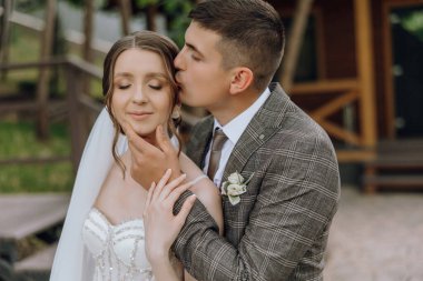 Bir gelin ve damat birbirlerinin boyunlarını öpüyor. Gelin beyaz bir elbise giyiyor ve damat da takım elbise giyiyor.