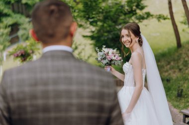 Bir gelin ve damat yeşil bir çalılığın önünde duruyor. Gelin bir buket çiçek tutuyor ve damat takım elbise giyiyor. Sahne mutlu ve kutlama dolu.