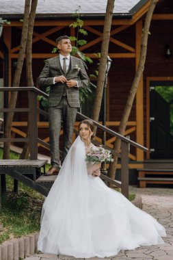 Gelin ve damat ahşap bir merdivende, gelin elinde buketle dikiliyorlar. Bu sahne muhtemelen bir düğün fotoğrafı, çünkü çift resmi giyinmiş.