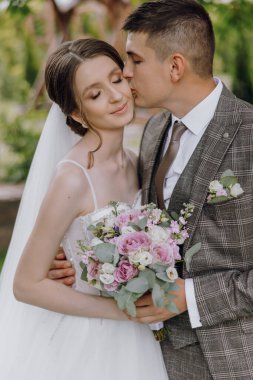 Bir gelin ve damat bir buket çiçeğin önünde birbirlerinin dudaklarını öpüyor. Gelin beyaz bir elbise giyiyor ve damat bir takım elbise giyiyor. Sahne romantik ve mutlu.