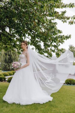 Beyaz elbiseli bir kadın bir ağacın önünde duruyor. Elinde bir buket çiçek ve duvak tutuyor. Sahne muhtemelen bir düğün ya da özel bir olay.