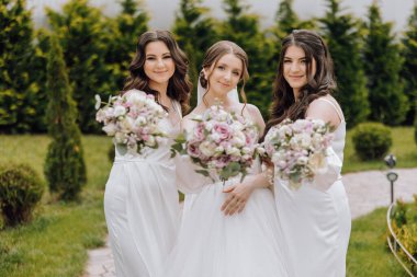Beyaz elbiseli üç kadın ellerinde çiçeklerle fotoğraf için poz veriyorlar. Sahne neşeli ve kutlayıcı, çünkü kadınlar büyük ihtimalle bir düğüne ya da diğer özel etkinliklere katılıyorlar.