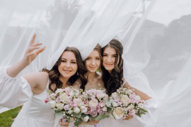 Üç kadın fotoğraf için poz veriyor, biri elinde bir buket çiçek tutuyor. Sahne beyaz perdeli ya da peçeli bir tarlada geçiyor. Sahne neşeli ve neşeli.