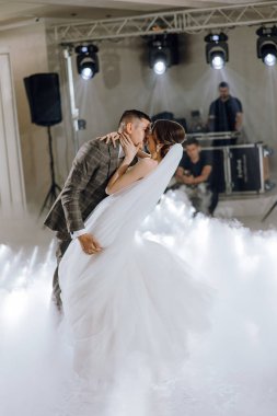 Bir gelin ve damat bir düğünde havada öpüşüyor. Gelin beyaz bir elbise giyiyor ve damat bir takım elbise giyiyor. Dans eden çiftin olduğu sahne canlı ve neşeli.