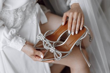 Bir kadın üzerinde fiyonk olan bir çift yüksek topuklu ayakkabı tutuyor. Ayakkabılar gümüş ve parıltılı, ve kadın beyaz bir elbise giyiyor. Zarafet ve incelik kavramı