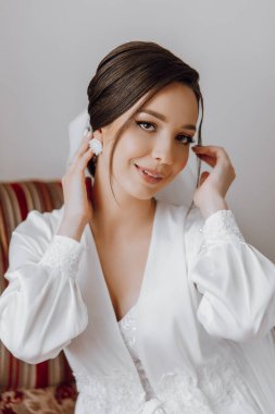 Beyaz elbiseli bir kadın kanepede oturuyor ve kulağına dokunuyor. Gülümsüyor ve özel bir durum için hazırlanıyor.