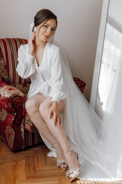 Beyaz elbiseli bir kadın kırmızı bir sandalyede oturuyor. Beyaz bir peçe takıyor ve elleri kalçasında.