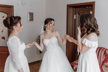 Üç kadın bir odada dikiliyor, biri gelinlik giyiyor. Hepsi gülümsüyor ve el ele tutuşuyorlar.