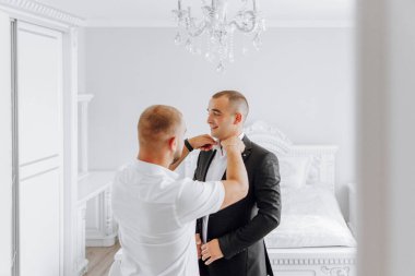 İki adam bir yatak odasında giyiniyor. Bir adam diğerine kravatını bağlamasında yardım ediyor.