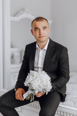 Takım elbiseli ve kravatlı bir adam elinde bir buket beyaz çiçek tutuyor. O bir yatakta oturuyor, kameraya bakıyor. Zarafet ve entelektüellik anlayışı, çünkü adam resmi giyinmiş.