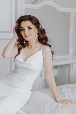 Beyaz bir elbise ve kolye giymiş bir kadın yatakta oturuyor. Uzun saçları var ve gülümsüyor.