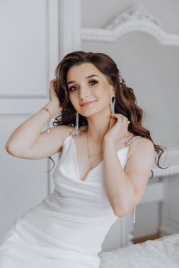 Beyaz elbiseli bir kadın yatakta oturuyor ve küpelerini takıyor. Yüzünde bir gülümseme var ve mutlu.