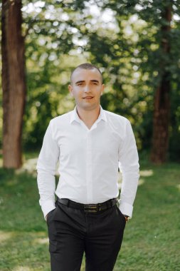 Beyaz gömlekli ve siyah pantolonlu bir adam çimen tarlasında duruyor. Gülümsüyor ve kameraya bakıyor. Rahatlama ve memnuniyet kavramı, adam dışarıda vakit geçirmekten zevk alıyor.