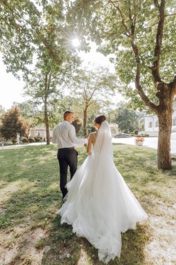 Bir gelin ve damat parkta birlikte yürüyorlar. Gelin beyaz bir elbise giyiyor ve damat beyaz bir gömlek giyiyor. Güneş parıldıyor, sıcak ve mutlu bir atmosfer yaratıyor.