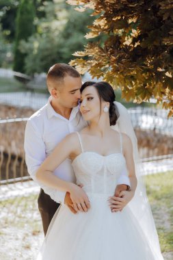 Bir gelin ve damat parkta kucaklaşıyor. Gelin beyaz bir elbise giyiyor ve damat beyaz bir gömlek giyiyor.