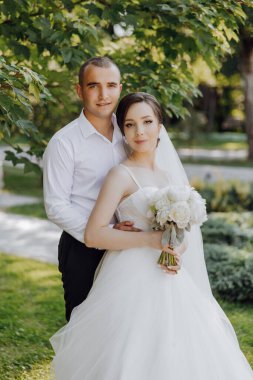 Bir gelin ve damat bir ağacın önünde fotoğraf için poz veriyorlar. Gelin elinde bir buket çiçek tutuyor.