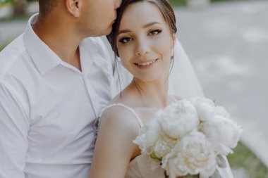 Gelin ve damat çimenli bir alanda oturuyor ve gelin elinde bir buket beyaz çiçek tutuyor. Çift gülümsüyor ve kameraya bakıyor, sıcak ve mutlu bir atmosfer yaratıyor.