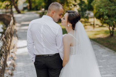 Bir gelin ve damat köprüde kameraya gülümsüyor. Gelin beyaz bir elbise giyiyor ve damat beyaz bir gömlek giyiyor. Sahne mutlu ve romantik.