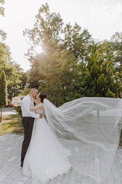 Bir gelin ve damat bir ağacın önünde öpüşüyor. Gelin beyaz bir elbise giyiyor ve damat beyaz bir gömlek giyiyor.