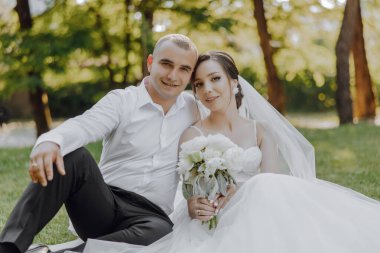 Bir gelin ve damat çimlerin üzerinde oturuyor, kameraya gülümsüyor. Gelin bir buket çiçek tutuyor ve damat onun elini tutuyor. Sahne mutlu ve romantik.