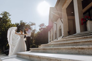 Gelin ve damat bir dizi basamağın üzerinde duruyorlar, gelin beyaz bir elbise tutuyor ve damat takım elbise giyiyor. Sahne bir kilisenin önünde geçiyor.