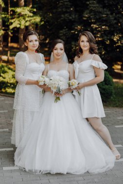 Üç kadın fotoğraf için poz veriyor, biri beyaz gelinlik giyiyor. Diğer iki kadın da beyaz elbiseler giyiyor ve ellerinde çiçekler var. Fotoğrafın mutlu bir tarafı var.
