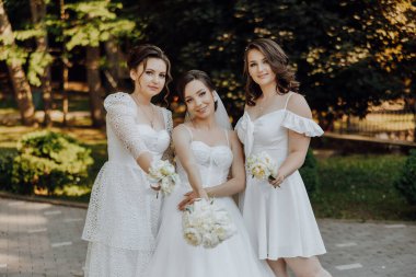 Beyaz elbiseli üç kadın fotoğraf için poz veriyor. Bir tanesi gelinlik giyiyor.