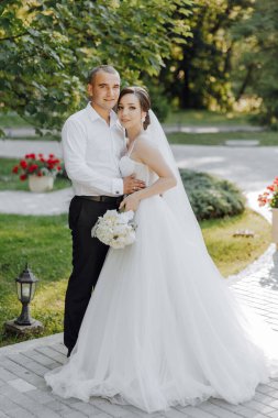 Bir gelin ve damat bir evin önünde fotoğraf için poz veriyorlar. Gelin beyaz bir elbise giyiyor ve damat beyaz bir gömlek giyiyor. Ellerinde buket ve buket tutuyorlar.
