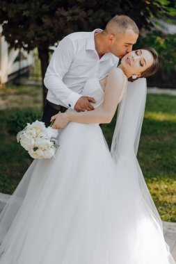 Bir gelin ve damat bahçede öpüşüyor. Gelin elinde bir buket beyaz çiçek tutuyor.