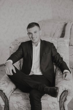 Takım elbiseli ve kravatlı bir adam sandalyede oturuyor. Gülümsüyor ve mutlu.