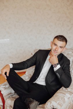 Takım elbiseli bir adam kanepede oturuyor ve kameraya bakıyor. O bir saat takıyor ve elinde kırmızı bir nesne var. Sofistike ve zarif kavramı