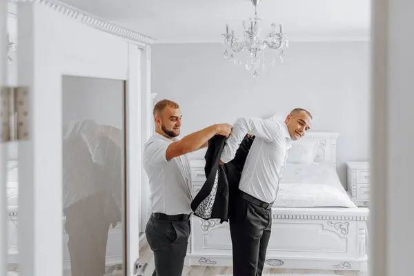 Two men are getting dressed in a bedroom. One man is adjusting the other man\'s jacket. Scene is casual and relaxed, as the men are getting ready for a day out