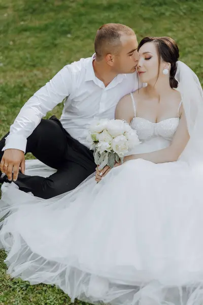 A bride and groom are sitting on the grass, the bride holding a bouquet of flowers. Scene is romantic and intimate