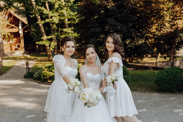 Beyaz elbiseli üç kadın bir evin önünde fotoğraf çektirmek için poz veriyor. Kadınlardan biri elinde bir buket çiçek tutuyor.