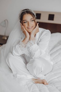 Beyaz elbiseli bir kadın elleri yüzü üzerinde bir yatakta oturuyor. Gülümsüyor ve mutlu.