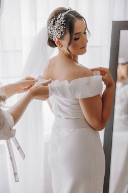 Bir kadın düğününe hazırlanıyor ve bir kadın duvağını düzeltmesine yardım ediyor. Gelin beyaz bir elbise ve beyaz bir peçe takıyor.