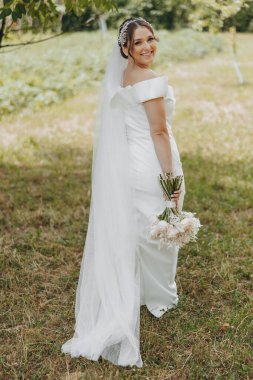 Beyaz bir duvak ve elinde çiçeklerle bir gelin tarlada duruyor. Gülümsüyor ve kameraya bakıyor.